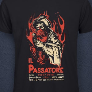 Könnte beinhalten: Ein schwarzes T-Shirt mit einem Vintage-Filmplakat-Design, das eine Person mit Kapuze zeigt, die eine Maske vor Flammen hält. Der Text auf dem Plakat lautet "IL PASSATORE" und enthält die Wörter "Basierend auf einer wahren Geschichte", "Stefano Pelion", "1851", "Avica Fornace" und "Forlimpopoli Theatre Production". Am unteren Rand des Shirts befindet sich ein kleines Etikett mit der Aufschrift "Cotone Organico" und "Antica For Nace Design".