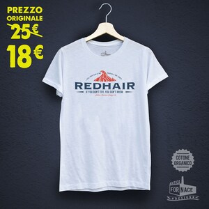 Könnte beinhalten: Weißes T-Shirt mit einem rot-blauen Logo, auf dem "Redhair" und "Wenn du es nicht versuchst, weißt du es nicht" steht. Das Logo enthält auch eine Welle mit Flammen. Das T-Shirt hängt an einem Kleiderbügel vor einem dunkelblauen Hintergrund.