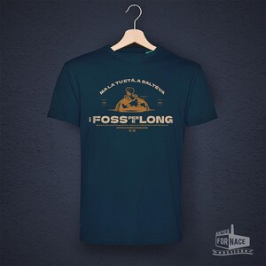 Könnte beinhalten: Ein tealfarbenes T-Shirt mit einem grafischen Design mit Text und einer Illustration. Der Text enthält "MALA TUETA, A SALTEVA" über einer Figur und die Worte "!FOSS PER LONG" darunter. Das Shirt hängt an einem Holzkleiderbügel.