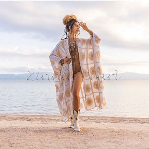 May include: A white crochet beach cover-up with gold embroidery. The cover-up has a long, flowing silhouette and a wide, open neckline. The model is wearing the cover-up over a brown swimsuit and white cowboy boots. The cover-up is photographed on a sandy beach with a body of water in the background.