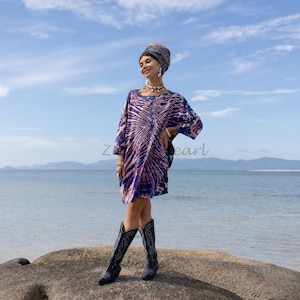May include: A short-sleeved dress featuring a blue and pink tie-dye pattern, paired with a dark patterned headwrap, a shell-style necklace, and black leather cowboy boots with white stitching details, worn in an outdoor coastal setting.