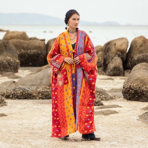 Puede incluir: Una larga y colorida bata con un diseño de patchwork. La bata presenta tonos vibrantes de rojo, naranja, amarillo y azul. La prenda tiene mangas largas y una silueta fluida. La bata es usada por una persona de pie en una playa arenosa.