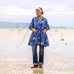 May include: A blue floral tunic with a V-neck and pockets, paired with dark wash jeans. The tunic features a colorful floral pattern in pink, blue, and white. The model is wearing a cream-colored headband and hoop earrings.