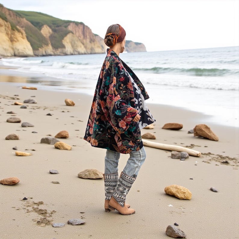 Side view of the Mei Velvet Kimono Duster in Nocturne Flora, showing its flowing silhouette and detailed floral velvet pattern. The model gazes toward the sea, wearing denim with tribal embroidery and neutral flats, her hair wrapped in a headscarf. The ocean waves and soft sky enhance the jacket’s romantic boho charm and handmade craftsmanship. Perfect depiction of ZinniaPearl’s slow fashion artistry.