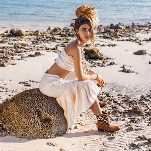 May include: A woman wearing a white crop top and a white skirt sits on a large rock. She is wearing brown boots with geometric patterns and has her hair pulled back in a bun. The background is a sandy beach with rocks and water.