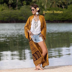 Op de afbeelding: Een vrouw draagt een mosterdgele kimono met een geometrisch patroon en een donkerbruine rand. De kimono is open en onthult een witte top en een spijkerbroek. De tekst "Saffron Sultan" is zichtbaar.
