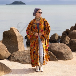 Full body front view of the open Mai velvet jacket styled with light wash denim jeans and white boots. The model stands with the coat open, revealing the fully lined interior and the generous width that fits up to a 72-inch bust. The "Golden Sunbird" print adds a pop of color with red and teal accents. A "Z" watermark is visible. This shot captures the versatility of the piece as both a robe and a stylish day coat.