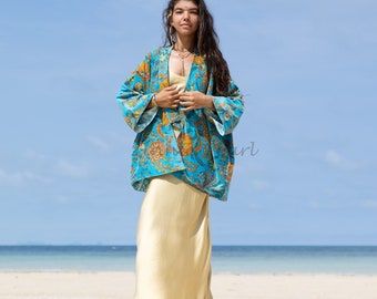 May include: A woman wearing a blue and gold floral patterned kimono jacket over a gold slip dress. She is standing on a sandy beach with a blue ocean in the background.