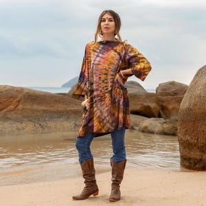 May include: A woman wearing a long, loose-fitting tie-dye tunic with a swirling pattern of orange, yellow, purple, and black. She is wearing blue jeans and brown cowboy boots. The tunic has a boat neck and three-quarter sleeves. The woman is standing on a sandy beach with large rocks in the background.