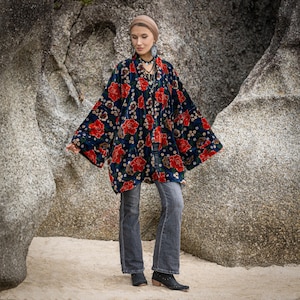 Könnte beinhalten: Eine marineblaue Kimono-Jacke mit einem auffälligen Blumenmuster aus roten und weißen Blüten. Die offen geschnittene Jacke hat weite Ärmel. Das Model trägt eine graue Jeans und schwarze Stiefel. Der Hintergrund ist eine natürliche Felsformation.