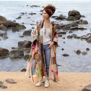 Op de afbeelding: Een vrouw draagt een lange, kleurrijke patchwork kimono met een wit topje en blauwe jeans. Ze staat op een strand met rotsen en water op de achtergrond. Ze draagt een turban op haar hoofd.