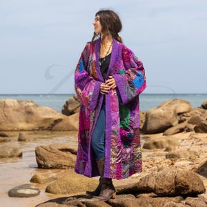 May include: A patchwork kimono in shades of purple, pink, and blue. The long, open-front robe features a quilted design with various fabric patterns. The kimono is worn over a black top and blue jeans, with brown boots.