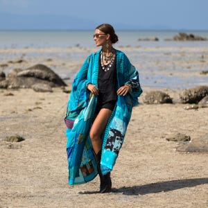 May include: A long, flowing patchwork kimono in various shades of blue, featuring a mix of floral and geometric patterns. The garment is worn over a black sleeveless top and black shorts, paired with black boots and a layered silver coin necklace.