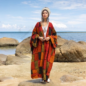 May include: A woman wearing a long, patchwork kimono with a variety of colors, including red, orange, green, and black. She is standing on a rocky beach with the ocean in the background.