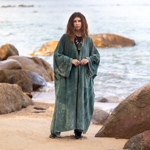 May include: A long, flowing, teal-colored velvet robe with wide sleeves. The robe is open in the front and worn over a black top. The person is standing on a sandy beach with rocks and the ocean in the background.