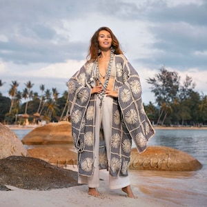 May include: A woman wearing a gray crochet kimono with white embroidery and white pants. She is standing on a beach with rocks and palm trees in the background.
