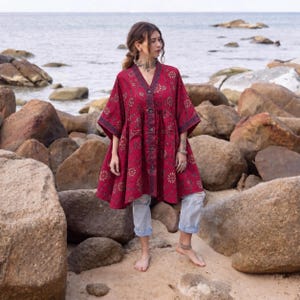 May include: A deep red tunic featuring a V-neckline, a front button placket, and short wide sleeves. The garment is decorated with a repeating floral pattern in a darker shade and is paired with light blue distressed denim jeans.