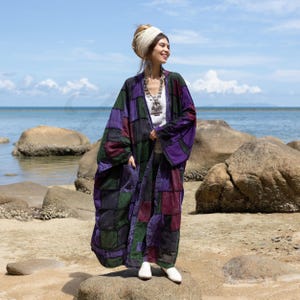 Könnte beinhalten: Eine Frau trägt eine lange, gepatchte Jacke im Kimono-Stil mit violetten, grünen und schwarzen Quadraten. Sie steht auf einem Felsen am Meer und trägt einen weißen Kopftuch und eine silberne Halskette.