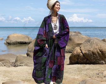 May include: A woman wearing a long, patchwork kimono-style jacket with purple, green, and black squares. She is standing on a rock by the ocean and wearing a white head wrap and a silver necklace.