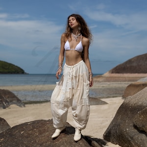 May include: A woman wearing a white bikini top and white, loose-fitting pants with a patchwork design. She is standing on a rock with the ocean in the background.