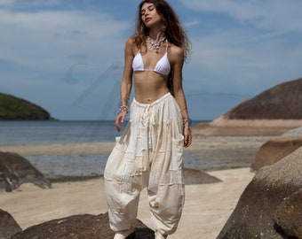 May include: A woman wearing a white bikini top and white, loose-fitting pants with a patchwork design. She is standing on a rock with the ocean in the background.