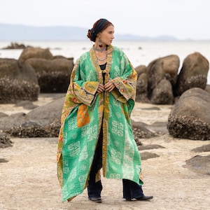 May include: A long, open-front green kimono with a floral pattern and colorful patchwork details. The kimono is worn over a black top and dark blue pants. The sleeves and edges are trimmed with patterned fabric. The background is a beach scene.