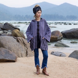 May include: A purple and black patterned hooded jacket with a black zipper and buttons. The jacket has two pockets on the front. The person wearing the jacket is standing on a beach with rocks in the background.