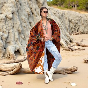 Model wearing the Amber Flora Harper Jade Velvet Kimono Robe by ZinniaPearl, standing on a sandy beach with driftwood and stone cliffs in the background. The plus size boho duster features burnt sienna velvet with jewel-toned florals in emerald, teal, and magenta, oversized wide sleeves, and deep front pockets. A handcrafted fall coat designed for holiday gatherings, bohemian festivals, and everyday luxe comfort, styled with distressed jeans and white boots.