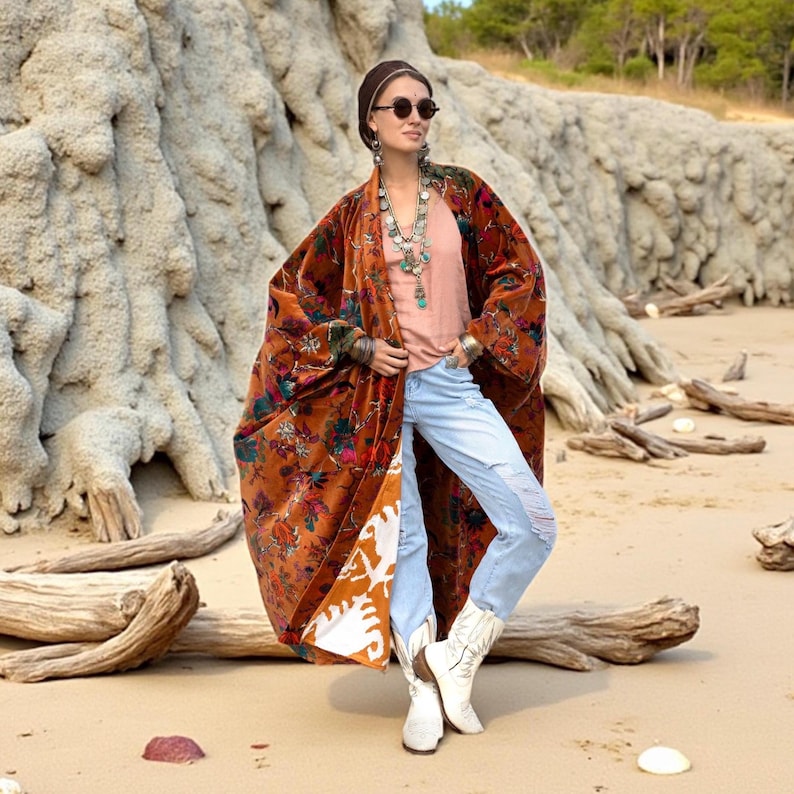 Model wearing the Amber Flora Harper Jade Velvet Kimono Robe by ZinniaPearl, standing on a sandy beach with driftwood and stone cliffs in the background. The plus size boho duster features burnt sienna velvet with jewel-toned florals in emerald, teal, and magenta, oversized wide sleeves, and deep front pockets. A handcrafted fall coat designed for holiday gatherings, bohemian festivals, and everyday luxe comfort, styled with distressed jeans and white boots.
