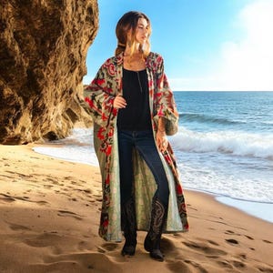 May include: A long, flowing, green and red patterned kimono with a black top and blue jeans. The person is standing on a sandy beach with the ocean in the background.