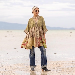 May include: A loose-fitting, button-down tunic featuring a patchwork design with various floral and abstract patterns in shades of olive green, brown, and cream, paired with blue denim jeans, a cream headband, and dark round sunglasses.