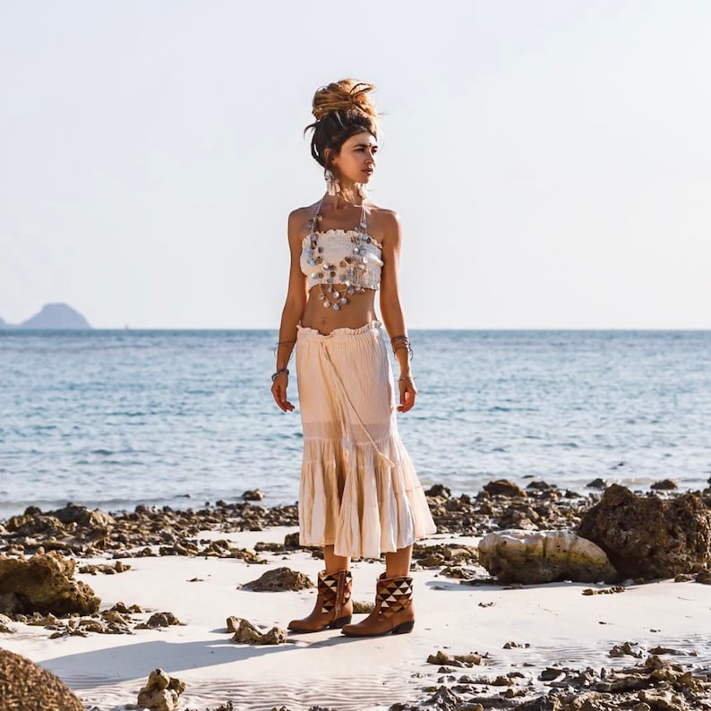 May include: A woman wearing a white crop top with a shell necklace and a white flowy skirt. She is standing on a sandy beach with the ocean in the background. She is wearing brown boots with a geometric pattern.