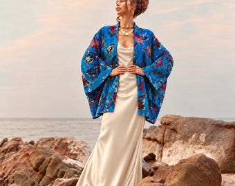 May include: A bright blue kimono-style jacket featuring a vibrant floral pattern in shades of pink, white, and green, worn over a floor-length, sleeveless, cream-colored satin slip dress with a scoop neckline and a fluid, straight-cut skirt.