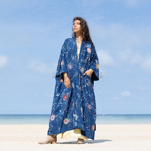 May include: A long, flowing blue kimono-style robe with a colorful, abstract pattern of circles and splatters. The robe has long sleeves and pockets. The model is wearing a yellow dress underneath. The background is a beach with the ocean and blue sky.