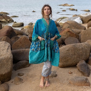 Peut inclure: Un vêtement de style kimono turquoise et sarcelle avec un motif floral sur la moitié inférieure et les manches. Le vêtement a un col en V et des poches. La personne se tient sur une plage de sable avec des rochers et l'océan en arrière-plan.