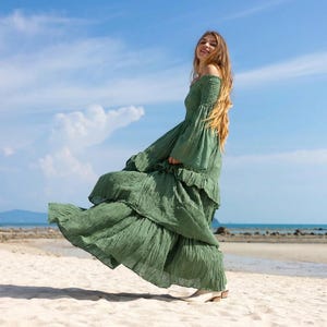 以下が含まれることがあります： オフショルダーの、オリーブグリーンの流れるようなドレス。ティアードフリルと長いベルスリーブが特徴です。 ドレスは、砂浜で着用されており、青い空と海を背景に、テクスチャード加工された生地で作られています。