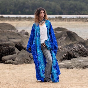 May include: A long, open-front kimono robe in shades of blue with a detailed floral and geometric pattern. The garment features wide sleeves and a flowing silhouette, worn over a plain white tank top and grey denim jeans with a layered necklace.