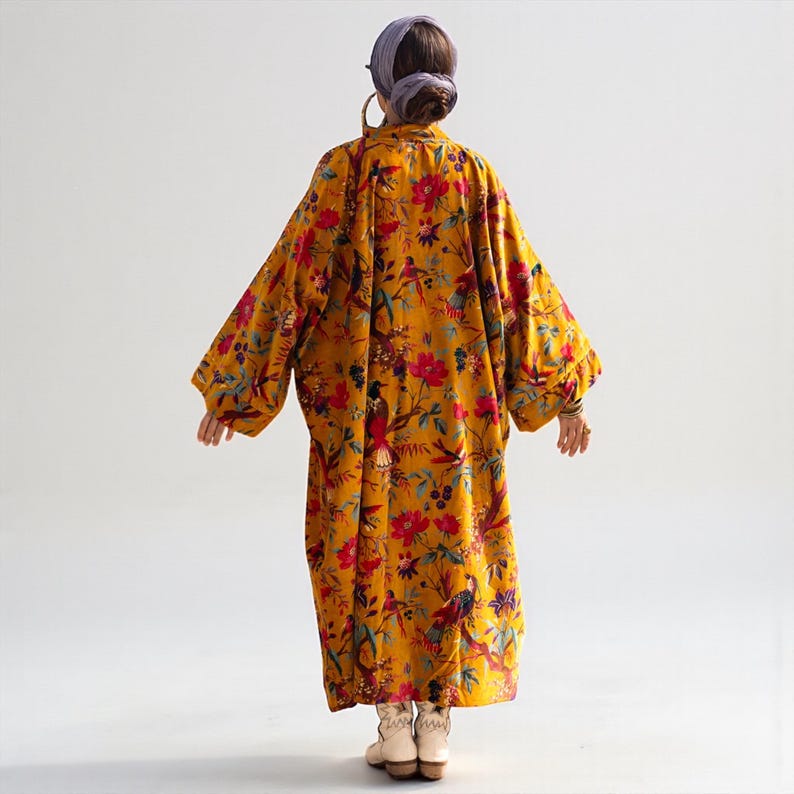 Back view of the Golden Sunbird velvet duster in mustard gold. The rich cotton velvet fabric features a vibrant print of colorful tropical birds and red floral blooms. The maxi-length kimono coat drapes loosely to the ankles, showcasing the wide, oversized fit and drop shoulders. Model wears a headwrap and white boots, highlighting the bohemian aesthetic.