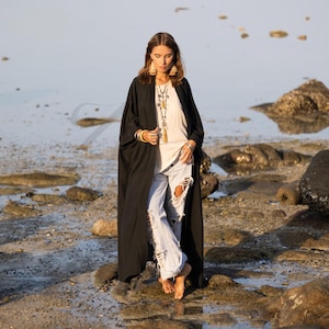 May include: A long, black open-front kimono-style robe. The robe is worn over a white top and distressed blue jeans. The model is standing on a rocky beach, with the water in the background. The robe is a flowing, lightweight fabric.