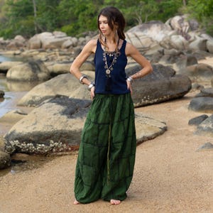 May include: A woman wearing a navy blue tank top and green wide-leg pants. The pants have a grid-like pattern. The woman is standing on a sandy beach with rocks and water in the background. She is wearing jewelry.