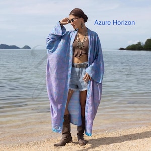 May include: A long, flowing kimono in shades of blue and purple, with the text "Azure Horizon" in blue. The kimono is worn open, revealing a brown top and denim shorts. The person is wearing brown boots and sunglasses, standing on a sandy beach.