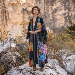Peut inclure: Un long kimono noir avec un motif patchwork coloré. Le kimono a une fermeture à nouer et est fait d'un tissu doux et fluide. Le mannequin porte le kimono avec un foulard et des bottes brunes.