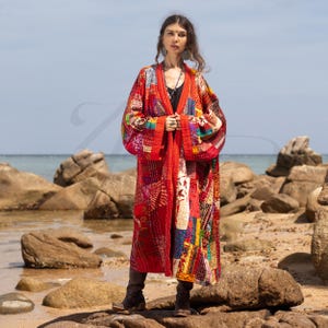May include: A long, red, patchwork kimono with colorful embroidery and a contrasting black trim. The kimono is worn by a person standing on a rocky beach.