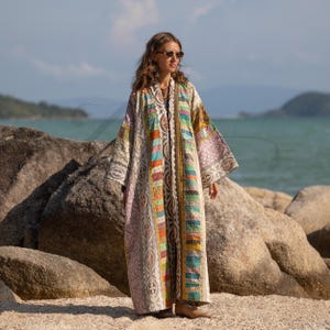 May include: A long, patchwork kimono-style robe with a variety of colorful rectangular patterns. The robe has a cream-colored base with accents of pink, blue, green, and yellow. The sleeves are wide, and the robe is worn outdoors.