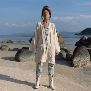 May include: A woman wearing a white linen jacket with a long, open front and a relaxed fit. She is standing on a sandy beach with large rocks in the background. She is wearing blue jeans and white cowboy boots.