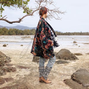 Side view of the Mei Velvet Kimono Duster in Nocturne Flora by ZinniaPearl, revealing its fluid silhouette and artisan-printed cotton lining. The model gazes toward the horizon under a coastal tree, highlighting the rich floral embroidery that glows against the dark velvet base. The composition blends craftsmanship, nature, and soulful femininity — a perfect expression of slow-fashion artistry.