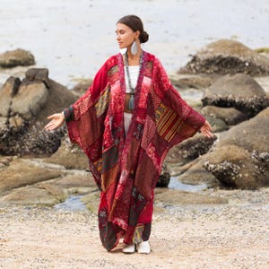 Peut inclure: Un kimono patchwork dans les tons de rouge, bordeaux et marron. Le kimono présente une variété de carrés de tissu à motifs cousus ensemble. Le vêtement est porté sur un haut et un pantalon blancs. Le modèle se tient sur une plage de sable avec des rochers en arrière-plan.