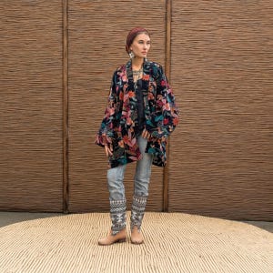 Model wearing the Mei Velvet Kimono Duster in Nocturne Flora by ZinniaPearl, standing against a woven rattan backdrop. The handcrafted black velvet jacket blooms with rich floral embroidery in shades of magenta, rust, jade, and copper. Styled with light denim and ethnic jewelry, this look captures ZinniaPearl’s signature bohemian luxury and artisanal craftsmanship — elegant, timeless, and inclusive.