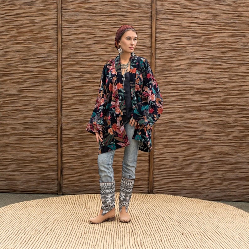 Model wearing the Mei Velvet Kimono Duster in Nocturne Flora by ZinniaPearl, standing against a woven rattan backdrop. The handcrafted black velvet jacket blooms with rich floral embroidery in shades of magenta, rust, jade, and copper. Styled with light denim and ethnic jewelry, this look captures ZinniaPearl’s signature bohemian luxury and artisanal craftsmanship — elegant, timeless, and inclusive.