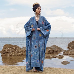 Puede incluir: Una larga bata de mezclilla azul con estampado de galaxia y bordados coloridos. La bata tiene un cuello ancho y abierto y mangas largas. La modelo lleva una cinta para la cabeza negra y joyas de plata.